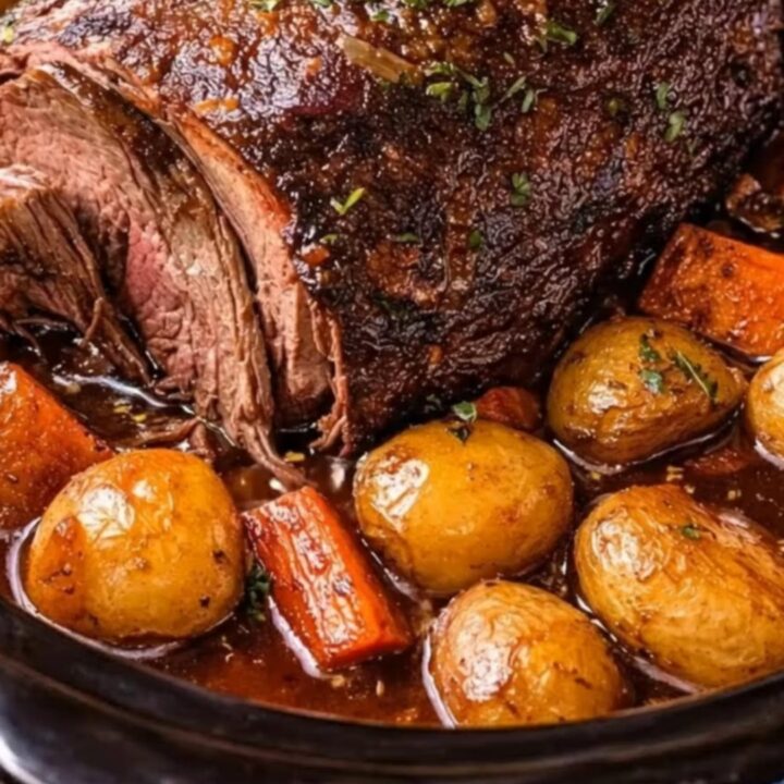 A glistening, herb-topped slow cooker pot roast resting in rich savory broth with tender baby potatoes and carrots.