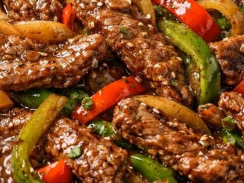 A black skillet filled with glossy pepper steak stir-fry garnished with sesame seeds and scallions.
