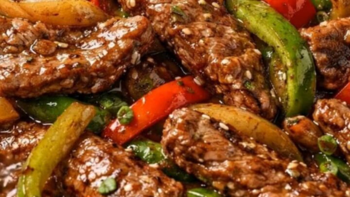 A black skillet filled with glossy pepper steak stir-fry garnished with sesame seeds and scallions.