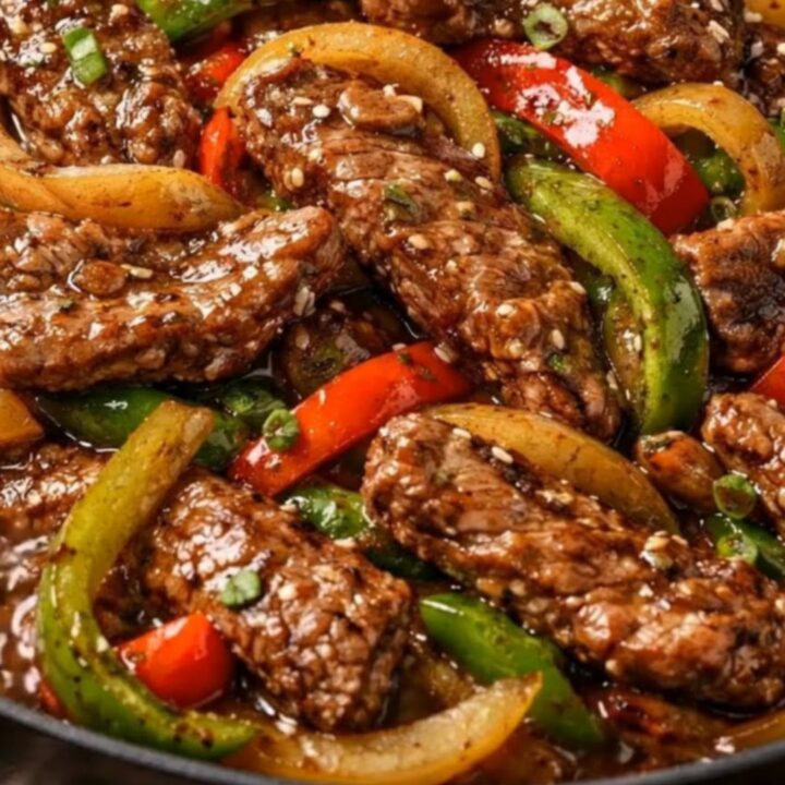 A black skillet filled with glossy pepper steak stir-fry garnished with sesame seeds and scallions.