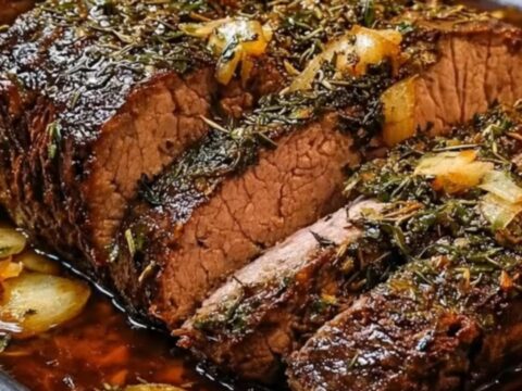 Sliced garlic herb roast beef in a dark roasting pan with caramelized onions and rich juices