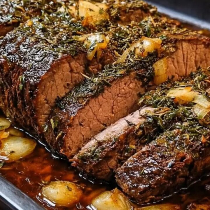 Sliced garlic herb roast beef in a dark roasting pan with caramelized onions and rich juices