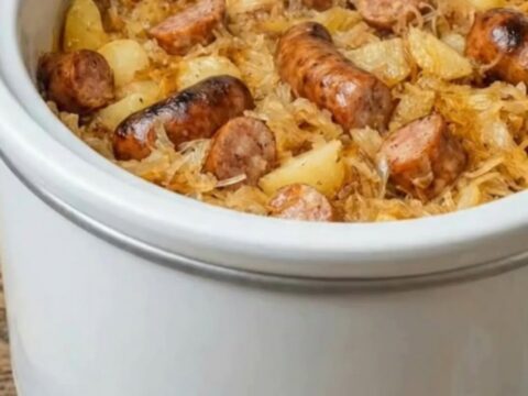 A white slow cooker filled with tender golden potatoes, juicy browned smoked kielbasa slices, and warm sauerkraut.