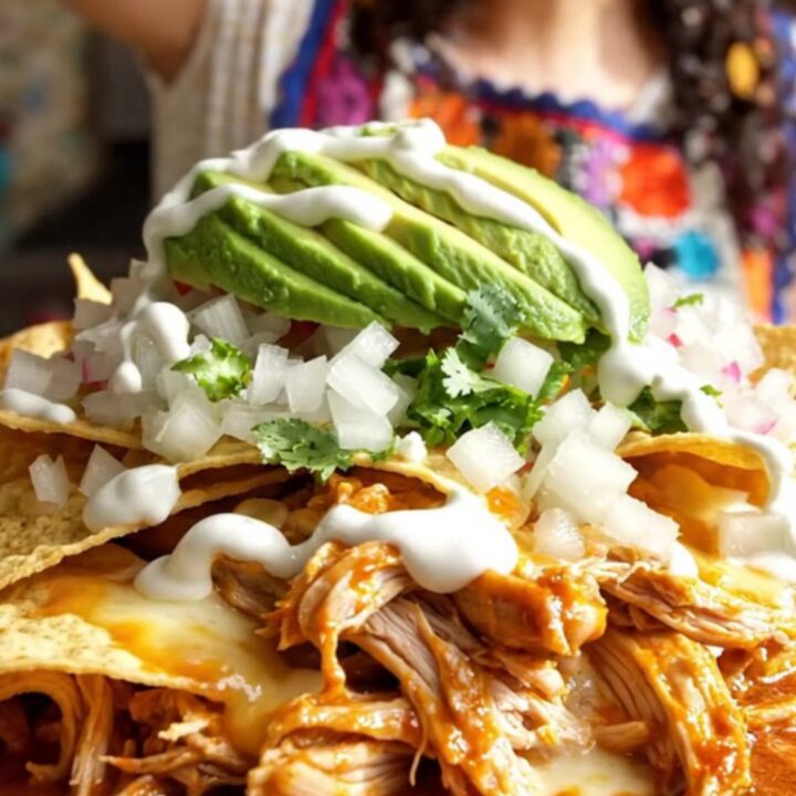 A steaming, rustic red ceramic slow cooker filled with cheesy, saucy chilaquiles topped with fresh avocado and cilantro.