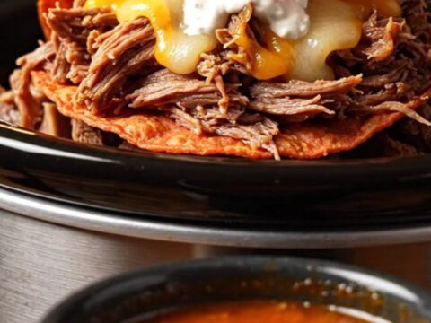 A close-up of a crispy quesabirria taco being dipped into a rich, red bowl of consommé.