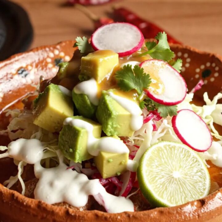 A steaming bowl of red pork posole topped with fresh cilantro, radish slices, and avocado in a rustic clay bowl.