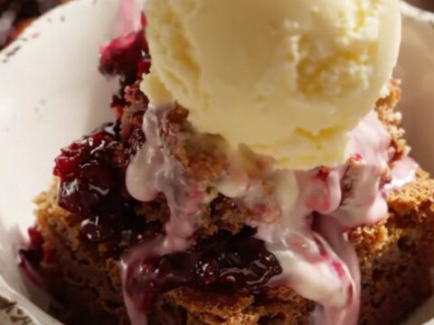 A close-up of a serving of warm blackberry bourbon dump cake topped with a melting scoop of butter pecan ice cream in a white