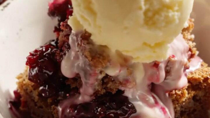A close-up of a serving of warm blackberry bourbon dump cake topped with a melting scoop of butter pecan ice cream in a white