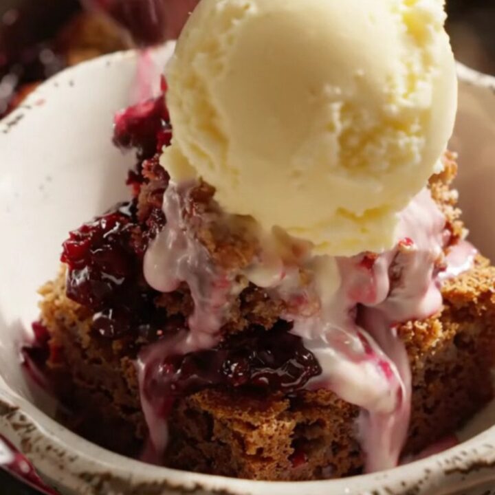 A close-up of a serving of warm blackberry bourbon dump cake topped with a melting scoop of butter pecan ice cream in a white