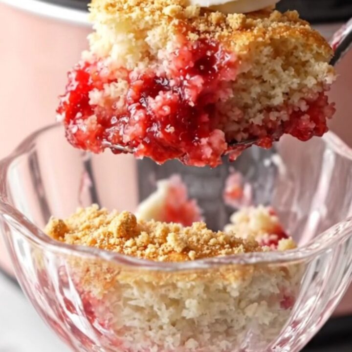 A close-up shot of a strawberry cheesecake dump cake in a blush-pink Crockpot with steam rising.