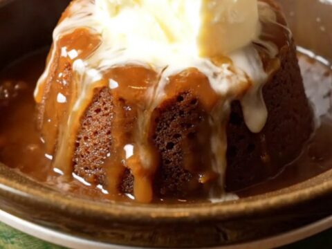 A steaming crockpot sticky toffee pudding topped with a dollop of clotted cream in a dark bowl.