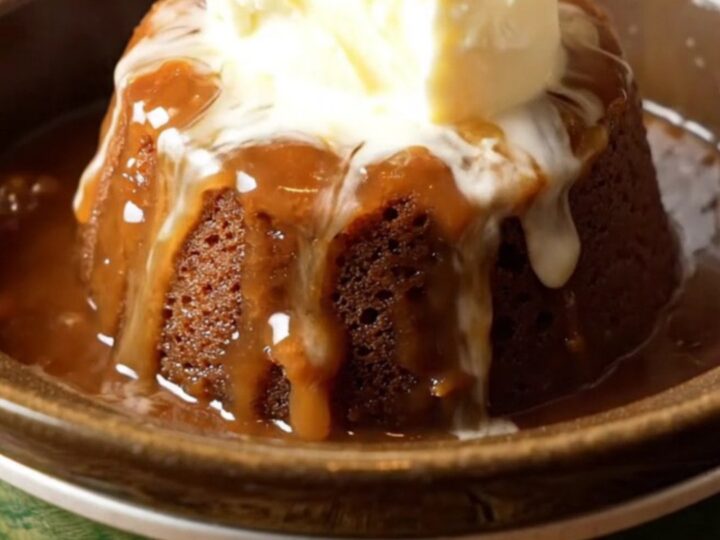 A steaming crockpot sticky toffee pudding topped with a dollop of clotted cream in a dark bowl.