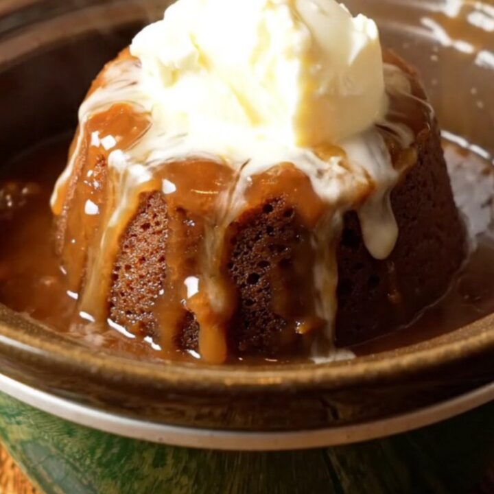 A steaming crockpot sticky toffee pudding topped with a dollop of clotted cream in a dark bowl.