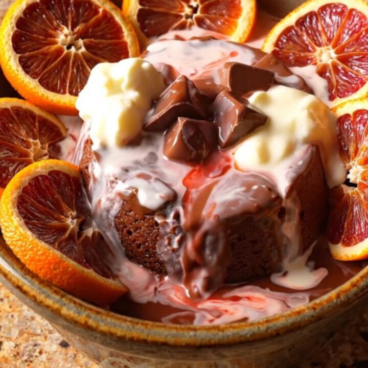 A close-up of dark chocolate blood orange lava pudding in a bowl, topped with candied citrus slices and cream.