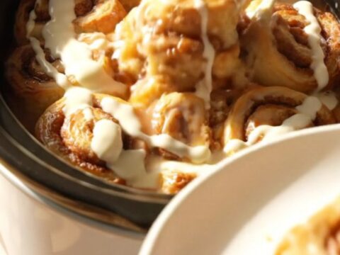 A warm, gooey cinnamon roll casserole served in a white ceramic slow cooker, topped with drizzled cream cheese icing.