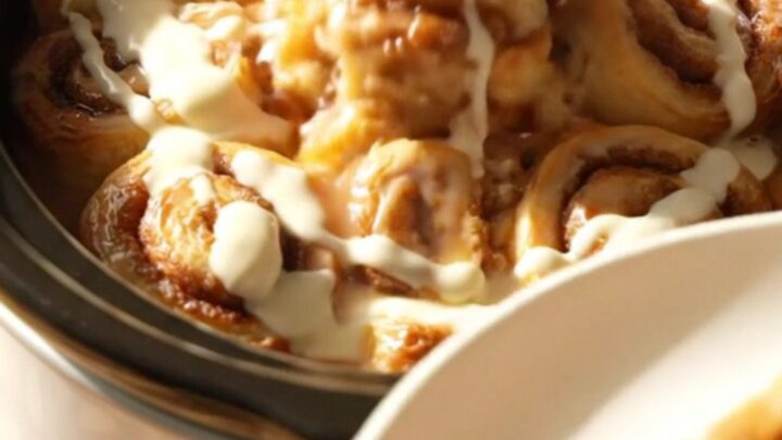 A warm, gooey cinnamon roll casserole served in a white ceramic slow cooker, topped with drizzled cream cheese icing.