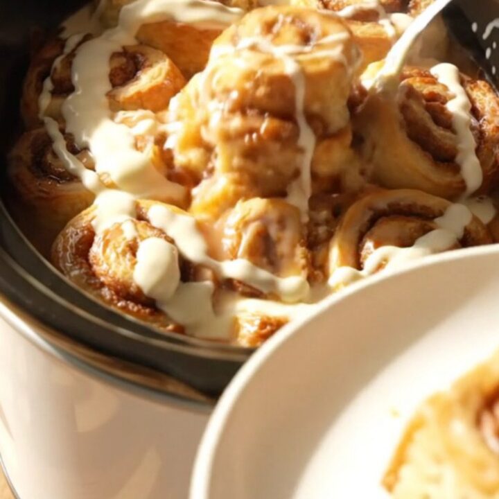 A warm, gooey cinnamon roll casserole served in a white ceramic slow cooker, topped with drizzled cream cheese icing.