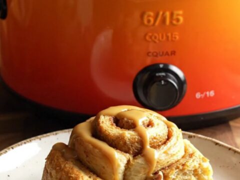 A golden, steaming pumpkin spice cinnamon roll casserole in a pumpkin-orange stoneware slow cooker.