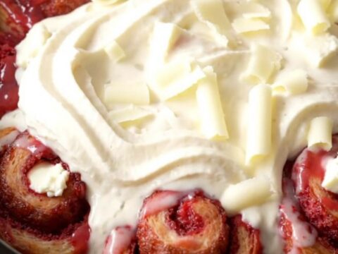 A vibrant red velvet cinnamon roll bake in a red ceramic slow cooker, topped with white icing and chocolate curls.