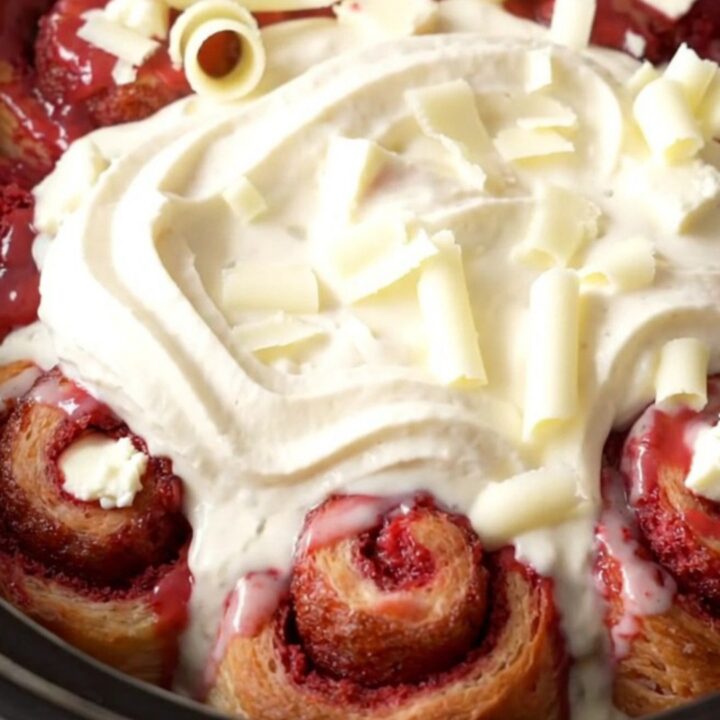 A vibrant red velvet cinnamon roll bake in a red ceramic slow cooker, topped with white icing and chocolate curls.