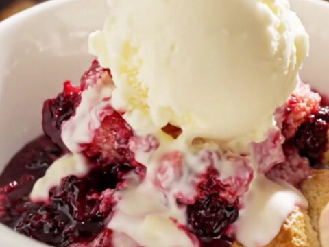 A close-up view of a spoon lifting a scoop of warm triple berry cobbler from a white ceramic slow cooker.