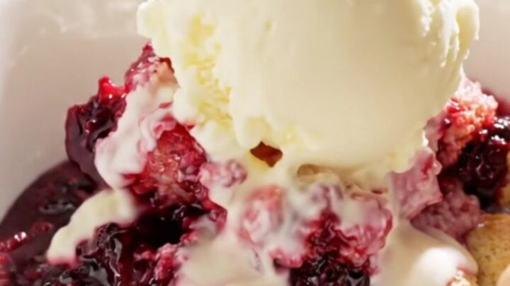 A close-up view of a spoon lifting a scoop of warm triple berry cobbler from a white ceramic slow cooker.