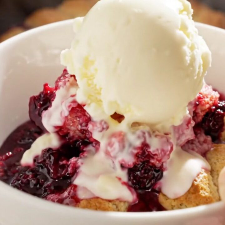 A close-up view of a spoon lifting a scoop of warm triple berry cobbler from a white ceramic slow cooker.