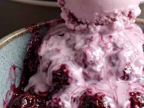 A close-up of a steaming bowl of purple boysenberry cobbler with a melting scoop of vanilla ice cream.