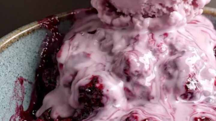 A close-up of a steaming bowl of purple boysenberry cobbler with a melting scoop of vanilla ice cream.