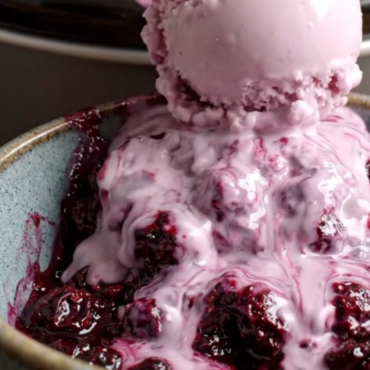 A close-up of a steaming bowl of purple boysenberry cobbler with a melting scoop of vanilla ice cream.