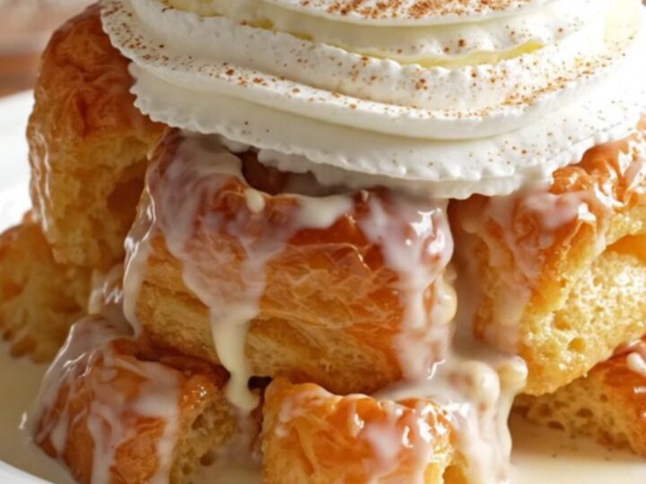 A warm, golden serving of tres leches brioche bread pudding topped with whipped cream and a cherry in a shallow bowl.