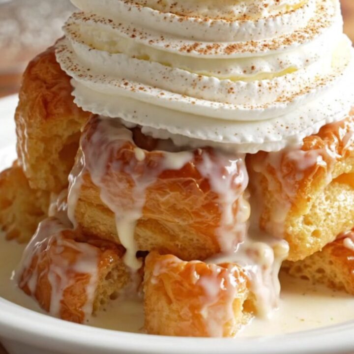 A warm, golden serving of tres leches brioche bread pudding topped with whipped cream and a cherry in a shallow bowl.