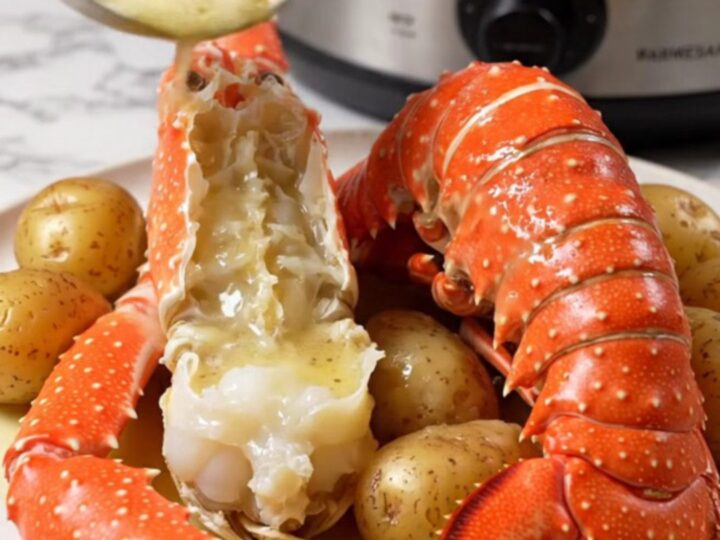 A close-up of split Alaskan king crab legs and lobster tails in a ceramic slow cooker with garlic butter.