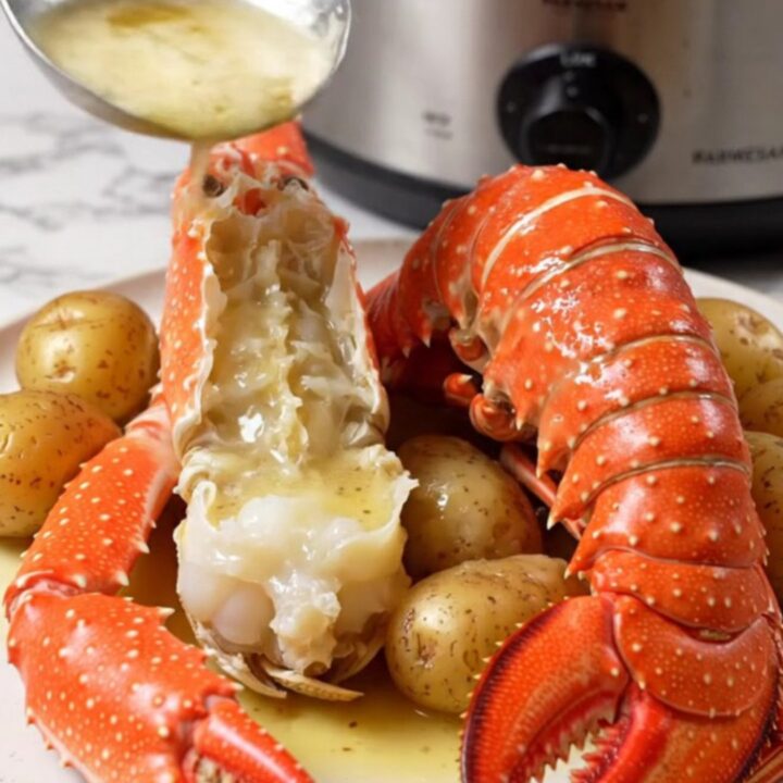 A close-up of split Alaskan king crab legs and lobster tails in a ceramic slow cooker with garlic butter.