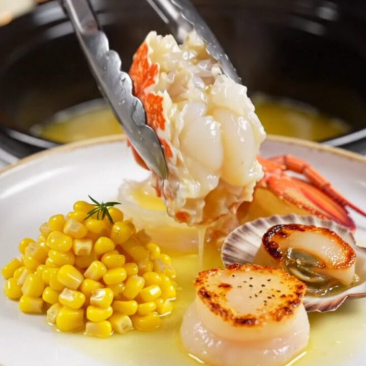 A close-up of butter-poached Maine lobster tails and sea scallops served on a warm plate with sweet corn.