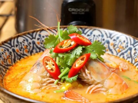 A close-up of a slow cooker filled with a vibrant red Thai Tom Yum seafood boil featuring prawns and mussels.