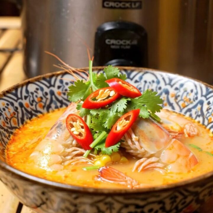 A close-up of a slow cooker filled with a vibrant red Thai Tom Yum seafood boil featuring prawns and mussels.