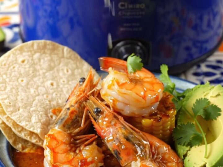 A close-up shot of fiery Camarones a la Diabla in a blue Crockpot, featuring large shrimp and charred corn.
