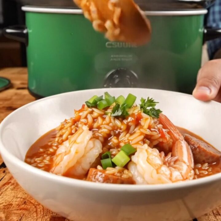 A close-up view of steaming jambalaya in a deep emerald ceramic Crockpot with a wooden lid handle.