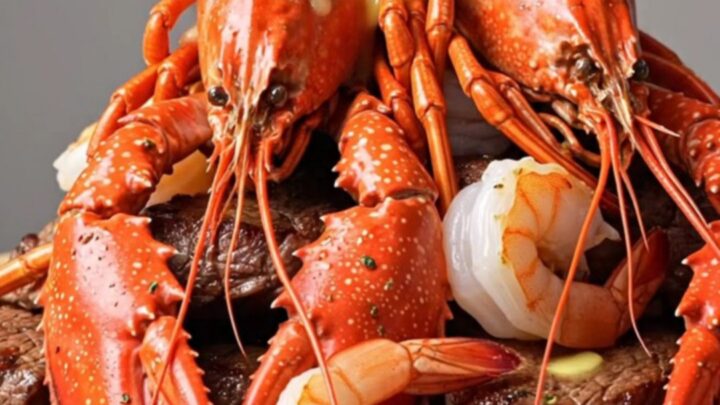 A rustic wooden board piled high with seared steak cubes, bright red lobster tails, and opaque shrimp