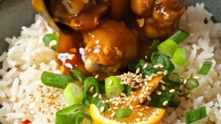 A bowl of sticky orange chicken served over fluffy white jasmine rice, garnished with sesame seeds and green scallions.