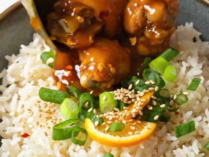 A bowl of sticky orange chicken served over fluffy white jasmine rice, garnished with sesame seeds and green scallions.