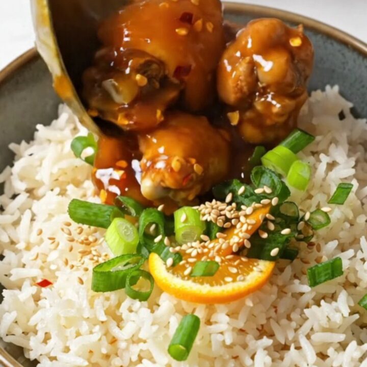 A bowl of sticky orange chicken served over fluffy white jasmine rice, garnished with sesame seeds and green scallions.
