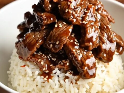 A bowl of tender, glossy Mongolian beef served over white rice with green onions and red pepper flakes.