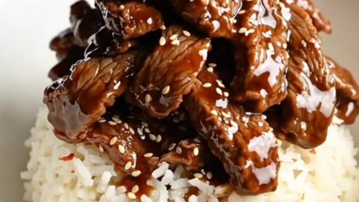 A bowl of tender, glossy Mongolian beef served over white rice with green onions and red pepper flakes.