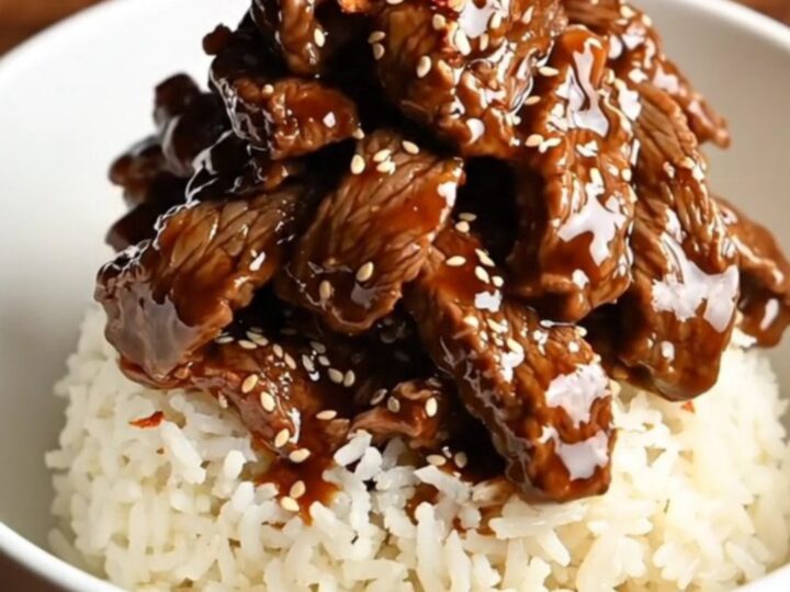 A bowl of tender, glossy Mongolian beef served over white rice with green onions and red pepper flakes.