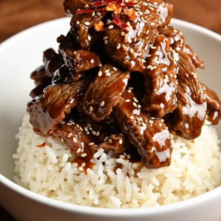 A bowl of tender, glossy Mongolian beef served over white rice with green onions and red pepper flakes.