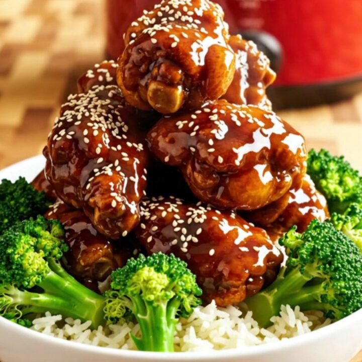 A close-up of General Tso's chicken in a crimson ceramic slow cooker, showing mahogany-glazed chicken with sesame seeds.