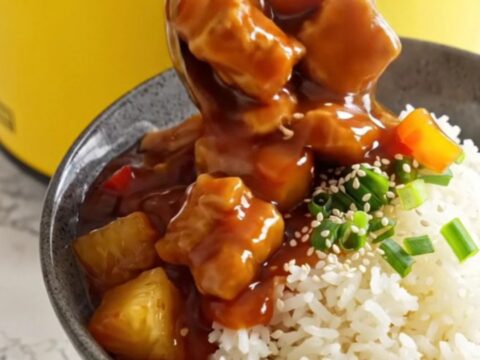 A ceramic slow cooker filled with sweet and sour pork, pineapple, and bell peppers in a glossy red sauce.