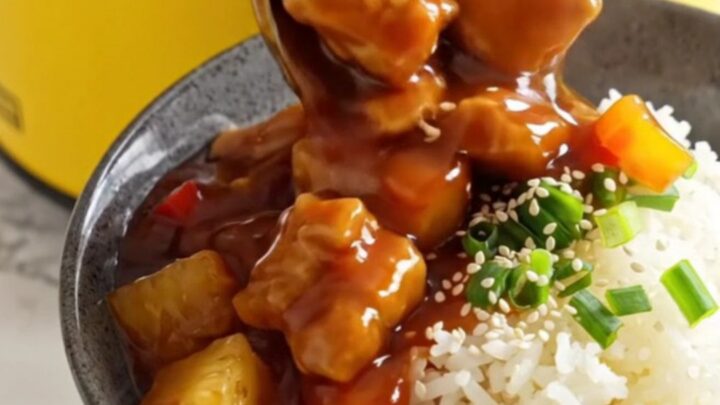 A ceramic slow cooker filled with sweet and sour pork, pineapple, and bell peppers in a glossy red sauce.